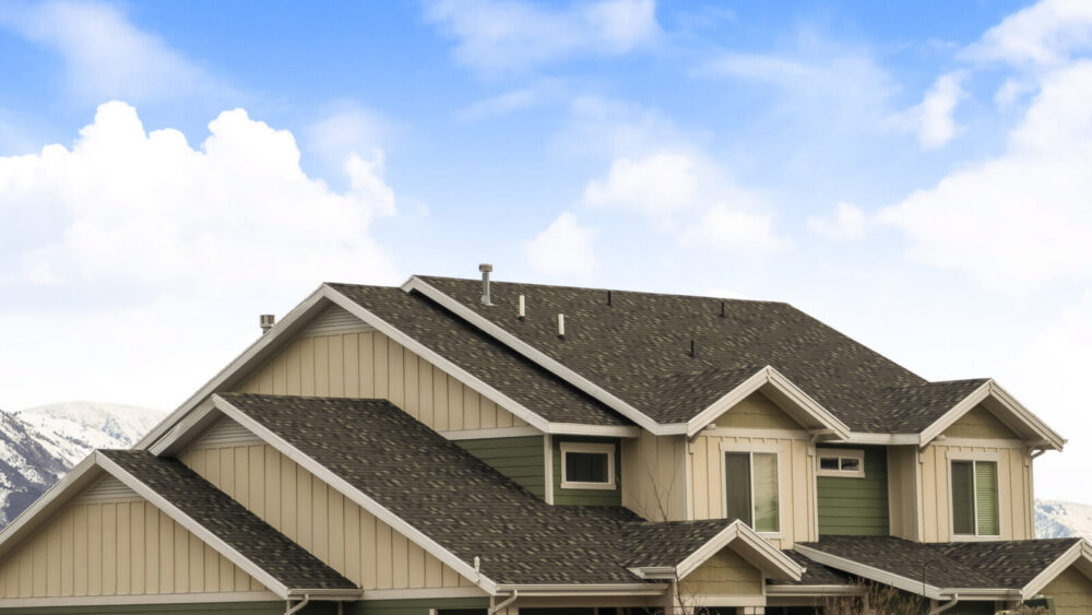 Panorama frame House exterior with view of the dark pitched roof against a cloudy blue sky. A mountain blanketed with snow can be seen behind the home.