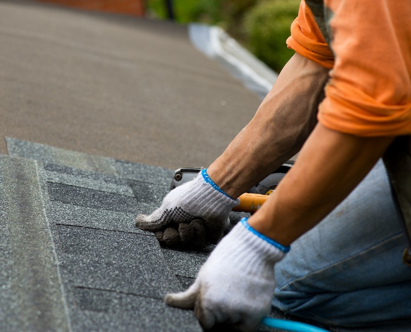 roofing contractor with white gloves and orange shirt applying roof shingles in the daylight.