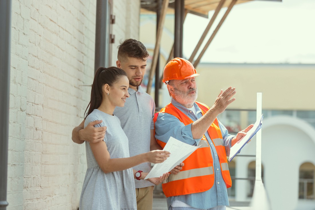 Foreman or achitect engineer shows future house, office or store design plans to a young couple. Meeting at the construction site to talk about facade appearance, interior decoration, home layout.